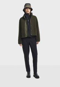 Žena v zelené fleecové bundě přes černou mikinu s kapucí, v černých kalhotách, černých botách a kostkovaném klobouku, stojící před jednobarevným pozadím.