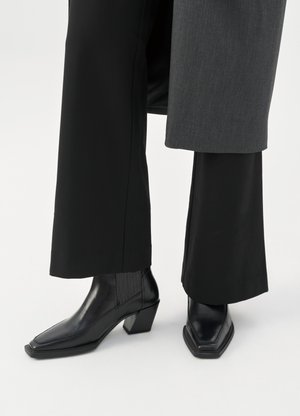 Schwarze Leder-Stiefeletten mit quadratischen Zehen und Blockabsätzen, kombiniert mit weiten schwarzen Hosen und einem grauen Mantel.