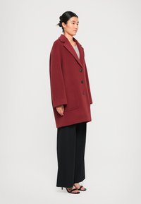 Manteau oversized en laine bordeaux avec de grands revers, trois boutons noirs et des poches latérales, porté sur un pantalon large noir et des sandales à talons.