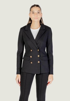 Giovane donna con capelli lunghi indossa un blazer nero doppiopetto con bottoni dorati e pantaloni aderenti abbinati, in piedi su uno sfondo chiaro.