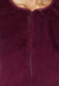 Vêtement en fausse fourrure bourgogne avec une surface douce et texturée, mettant en avant une fermeture centrale et un col arrondi.