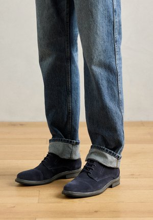 Marineblaue Wildleder-Stiefeletten mit runder Spitze, geschnürter Vorderseite und kontrastierendem Nähen, kombiniert mit leicht blauen Jeans mit umgeschlagenem Saum.