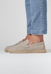 Beige suède loafers met een gestikt ontwerp, voorzien van een decoratieve klep en een gevlochten jute zool, gecombineerd met lichtblauwe cropped jeans.