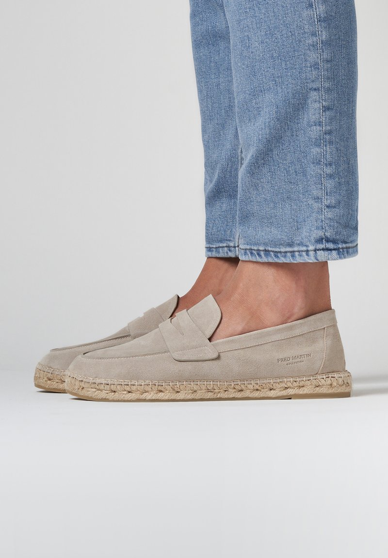 Beige suède loafers met een gestikt ontwerp, voorzien van een decoratieve klep en een gevlochten jute zool, gecombineerd met lichtblauwe cropped jeans.