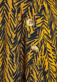 Tessuto giallo con un motivo a zig-zag nero; presenta due grandi bottoni; la consistenza appare leggera e tessuta.