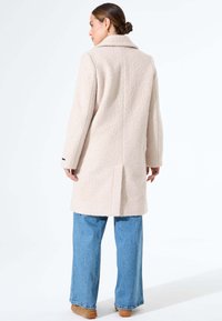 Lichtbeige textuurjas met een kraag, rugventilatie en lange mouwen, gecombineerd met wijdvallende blauwe denim jeans en tan-kleurige schoenen.