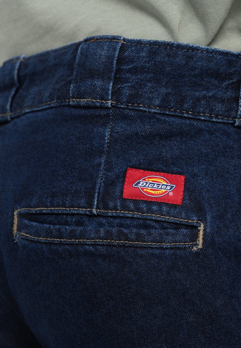 Vaqueros de mezclilla en azul oscuro con una textura suave. Incluye características como una cinturilla, bolsillos traseros, y una etiqueta de logo roja de Dickies cosida.