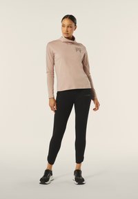 Maglietta rosa chiaro a maniche lunghe con collo drappeggiato e logo nero, abbinata a leggings neri aderenti, accentuata da sneakers nere sportive.