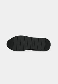 Musta kuminen tossun pohja vaakasuorilla uurteilla ja "Calvin Klein" -logo painettuna keskelle.