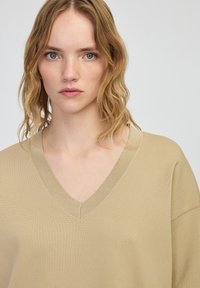 Beżowy sweter w splotach z dekoltem w serek. Charakteryzuje się luźnym krojem i szerokimi rękawami, prezentując prosty i nowoczesny design.