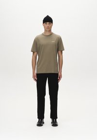 Olijfgroene t-shirt met korte mouwen en een logo op de borst, gecombineerd met zwarte slim-fit broek en zwarte schoenen. Model staat tegen een witte achtergrond.