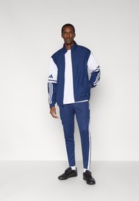 Giacca sportiva blu navy e pantaloni coordinati con strisce bianche, realizzati in tessuto liscio. Sneaker nere completano il look atletico.