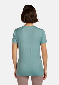 Mujer con cabello corto castaño que lleva una camiseta de manga corta color teal y pantalones marrón claro, vista desde la espalda contra un fondo liso.