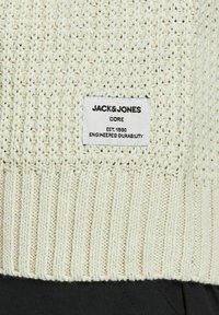 Pull en maille couleur crème avec ourlet à côtes et une étiquette rectangulaire blanche indiquant « JACK & JONES CORE EST. 1990 ENGINEERED DURABILITY. »