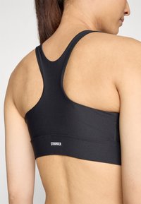 Črn športni modrček z zasnovo racerback, izdelan iz gladkega materiala. Ima majhen bel logotip "STRONGER" na spodnjem traku.