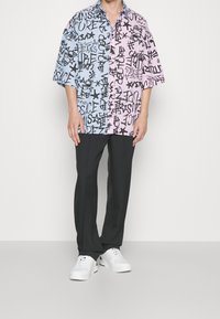 Camisa de manga corta partida en azul y rosa, con texto y diseños en estilo graffiti en negro, llevada con pantalones negros y zapatillas blancas.