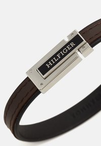 Brown leather bracelet featuring a silver-tone rectangular clasp with a black panel inscribed with "HILFIGER." Smooth texture; minimalistic design.