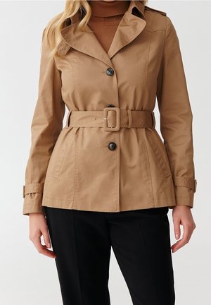 Kobieta ubrana w beżowy płaszcz trencz z paskiem i czarnymi guzikami, nałożony na brązowy golf, w połączeniu z czarnymi spodniami na jednolitym tle.