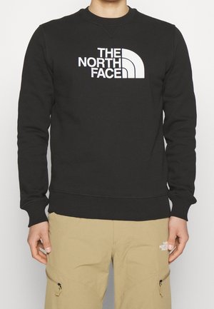 Person, die einen schwarzen Sweatshirt mit weißem "The North Face"-Logo und beige Hose mit kleinem weißem Logo am linken Oberschenkel trägt.