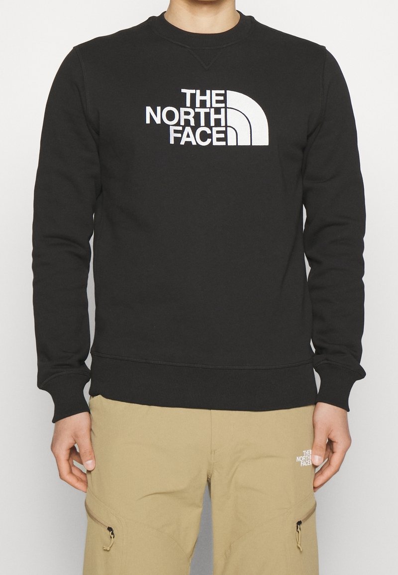 Personne portant un sweat-shirt noir avec le logo blanc "The North Face" et un pantalon beige avec un petit logo blanc sur la cuisse gauche.