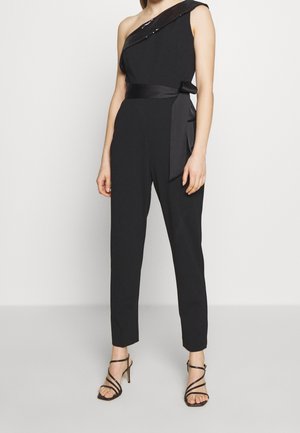Femme portant une combinaison noire une épaule avec une ceinture en satin à la taille et des sandales noires à talons hauts à brides sur un fond uni.