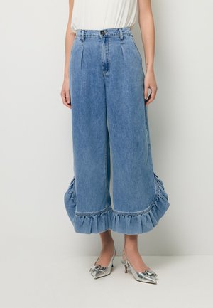 Kobieta ubrana w szerokie dżinsy o kroju dzwon z falbanami na nogawkach i srebrne szpiczaste szpilki, zestawione z białą bluzką.