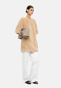 Kobieta w beżowej, puszystej, oversize'owej bluzie, białych szerokich spodniach, czarnych sandałach, trzymająca dużą, złożoną szaro-clutch za tłem w jednolitym kolorze.