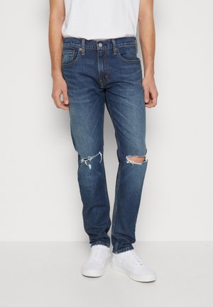 Jeans de mezclilla de un azul oscuro con un corte ajustado, que presentan desgastes en las rodillas y un diseño clásico de cinco bolsillos. Combinados con zapatillas blancas.