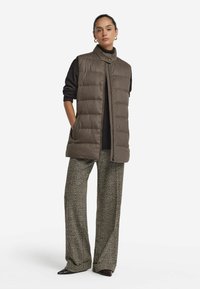 Puffig ärmlös väst i mörkbrunt tyg med hög krage, med två sidofickor och dragkedja framtill, matchad med mönstrade vida byxor.