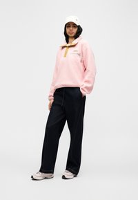 Ροζ φλις πουλόβερ με ψηλό γιακά και τσέπη με φερμουάρ· συνδυασμένο με σκούρο τζιν παντελόνι και ανοιχτόχρωμα αθλητικά παπούτσια. Το λευκό καπέλο ολοκληρώνει το σύνολο.