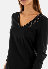 Maglietta nera a maniche lunghe con scollo a V, caratterizzata da dettagli metallici lungo il collo e un testo decorativo nella parte superiore sinistra.