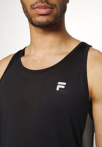 Homme portant un maillot de sport noir sans manches avec un logo blanc en forme de F sur la poitrine gauche, montrant son cou, ses lèvres et une partie de son visage.