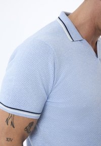 Ljusblå polo t-shirt med texturerad tyg, med en kontrasterande krage med marinblå och vita ränder. Korta ärmar, figursydd design.