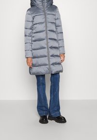 Persona che indossa un cappotto imbottito grigio lucido con cappuccio, jeans blu di denim e scarpe nere robuste, in piedi contro uno sfondo chiaro uniforme.