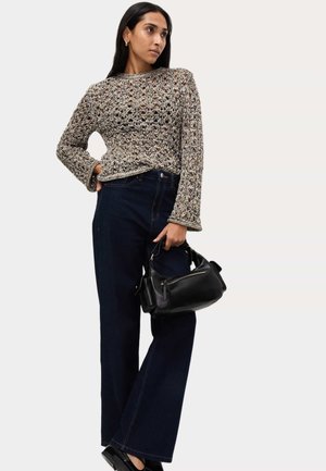 Donna che indossa un maglione beige all'uncinetto e jeans scuri a gamba larga, che tiene una borsa in pelle nera, posando con una mano sui fianchi.