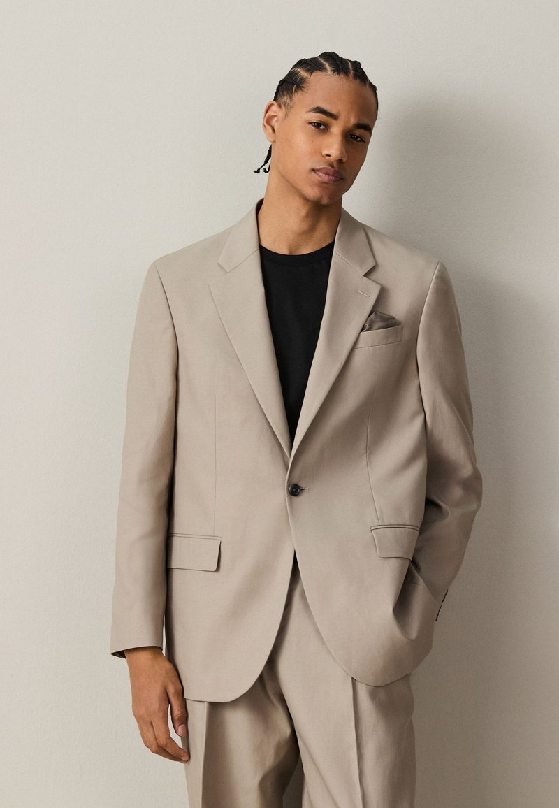 Young man with braided hair wearing a beige suit jacket and matching pants over a black shirt, standing with one hand in pocket.