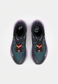 Une paire de chaussures de course vert foncé et noires avec des accents orange et des semelles violettes, vue de dessus sur un fond blanc.