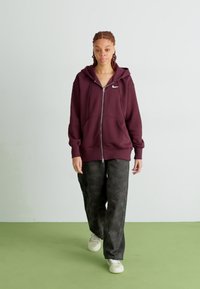 Sweatshirt marrom com fecho e bolsos canguru e pequeno logo, combinado com calças largas de padrão escuro e sapatilhas brancas sobre um chão verde.