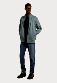 Jasnoniebieska kurtka zapinana na zamek z wysokim kołnierzem, połączona z czarną koszulą, niebieskimi dżinsami i czarnymi masywnymi butami. Gładka faktura i luźny krój.