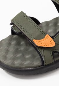 Sandalo verde oliva con tomaia testurizzata, cinturino in velcro, dettaglio arancione e suola in gomma nera. Presenta una vestibilità regolabile e un design aperto.