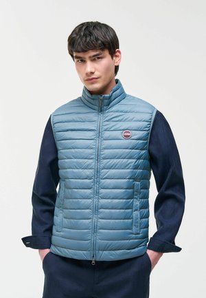 Jonge man met een lichtblauw gewatteerd mouwloos jack over een donker shirt met lange mouwen, staand met handen in de zakken.