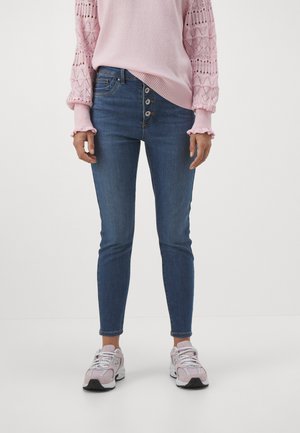 Skinny jeans in dark blue denim with five-button front closure, paired with a light pink sweater featuring textured sleeves and ruffled cuffs.