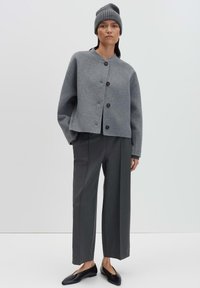 Donna che indossa un berretto grigio, una giacca grigia oversized con bottoni, pantaloni larghi color antracite e scarpe flat nere, in piedi con una mano in tasca.