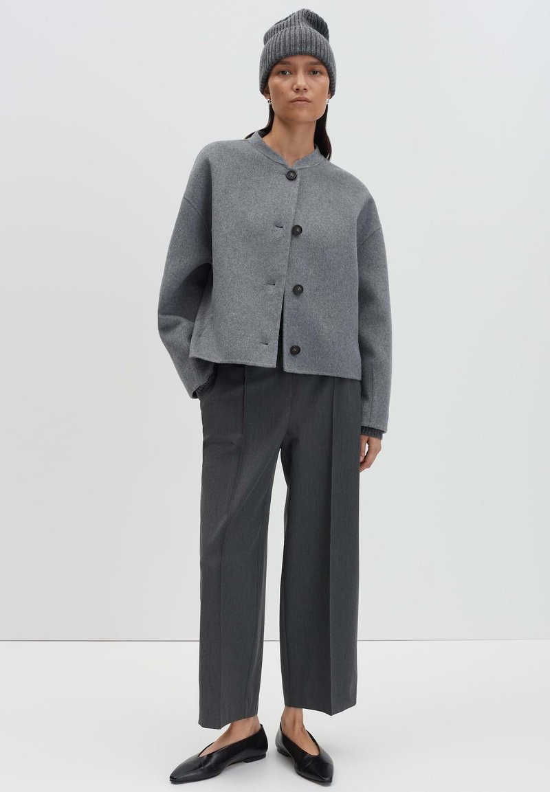 Donna che indossa un berretto grigio, una giacca grigia oversized con bottoni, pantaloni larghi color antracite e scarpe flat nere, in piedi con una mano in tasca.