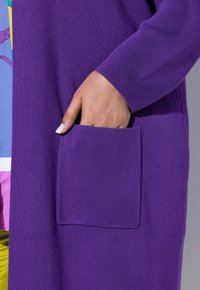 Paarse gebreide cardigan met een gestructureerde afwerking, met een voorzak en geribde manchetten, die gedeeltelijk een kleurrijke top bedekt.
