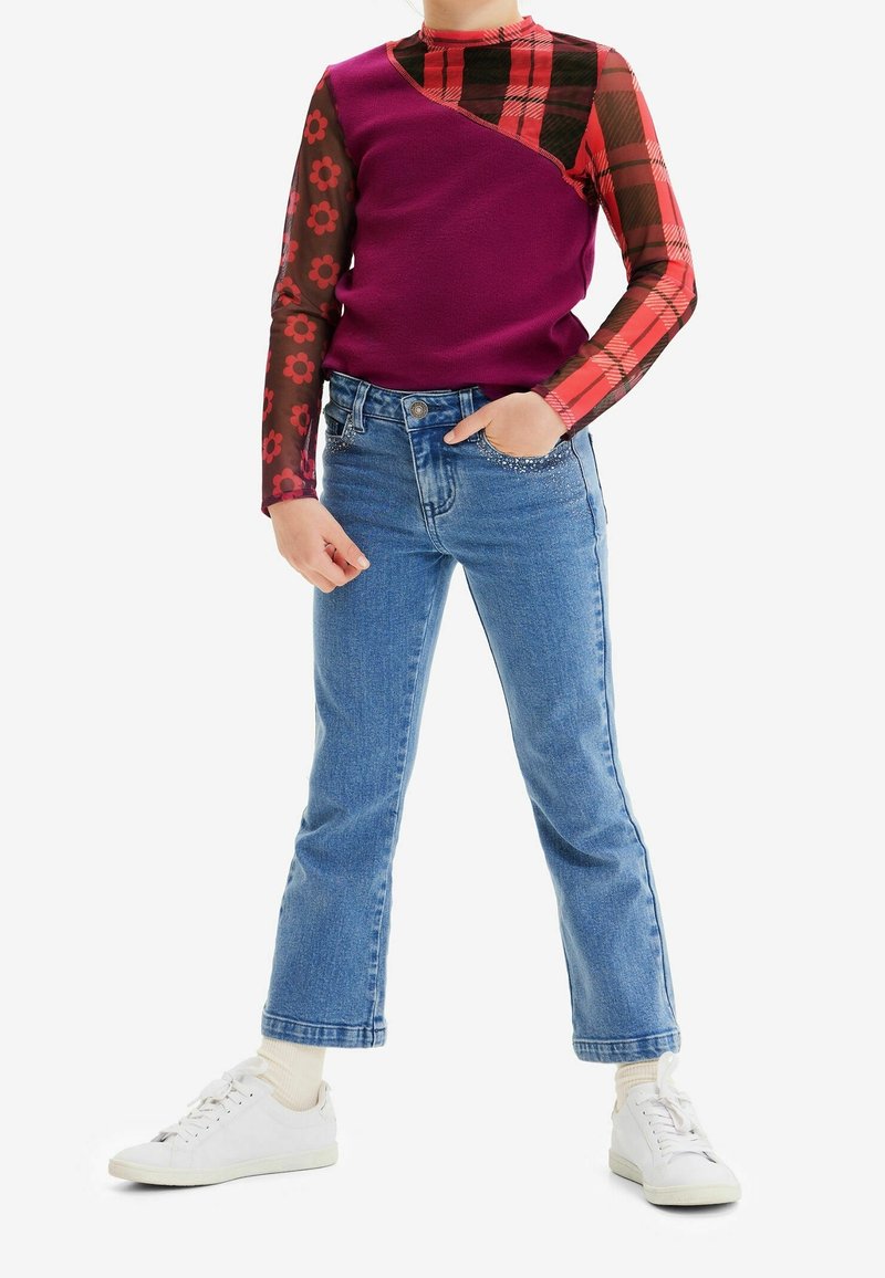 Buntes Langarmshirt in Lila mit rot karierten und gemusterten Ärmeln, kombiniert mit hellblauen Jeans und weißen Sneakern.