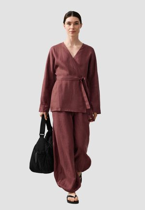 Kobieta idąca, ubrana w luźną bordową bluzkę z kopertowym dekoltem i spodnie, trzyma czarną torbę, ma czarne sandały, neutralne tło.