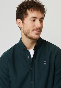 Donkergroene button-up shirt van een zachte stof, met een klein wit geborduurd boomlogo op de linkerborst.