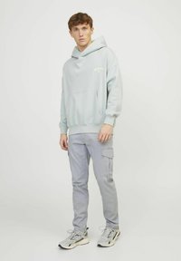 Junger Mann in einem hellblauen Hoodie, grauen Cargo-Hosen und Turnschuhen, der vor einem schlichten weißen Hintergrund steht.