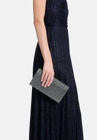 Pochette métallique grise avec un motif géométrique texturé tenue dans une main. Le design est élégant et rectangulaire, complétant une robe bleu foncé.